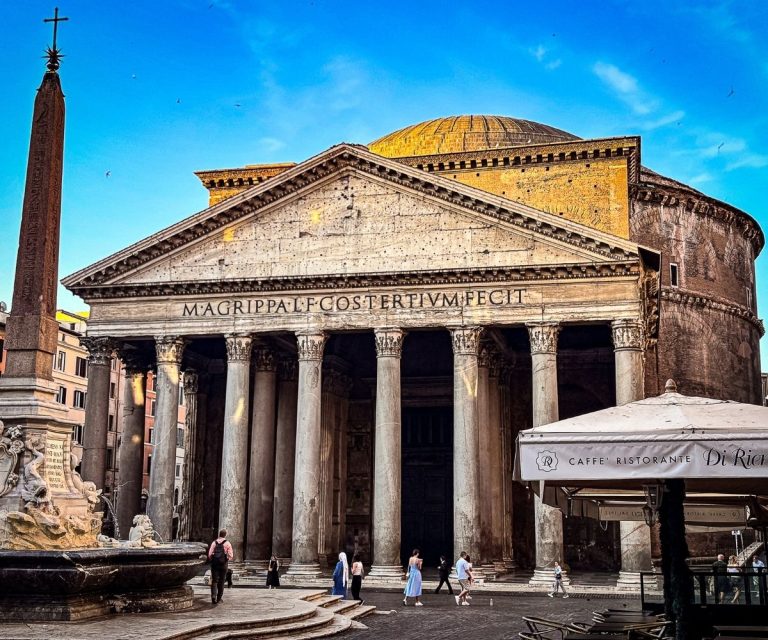 People walking in front of the Pantheon in Rome just after sunrise, with soft light hitting the ancient columns, dome, and nearby fountain on a clear blue-sky morning.