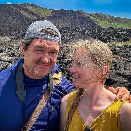 A smiling couple poses for a selfie during a sunny hike across a rugged volcanic landscape in Sicily. The man wears a backwards cap and camera strap, while the woman in a yellow top looks at him with a warm smile. Behind them, black lava rocks stretch toward green hills under a bright blue sky.