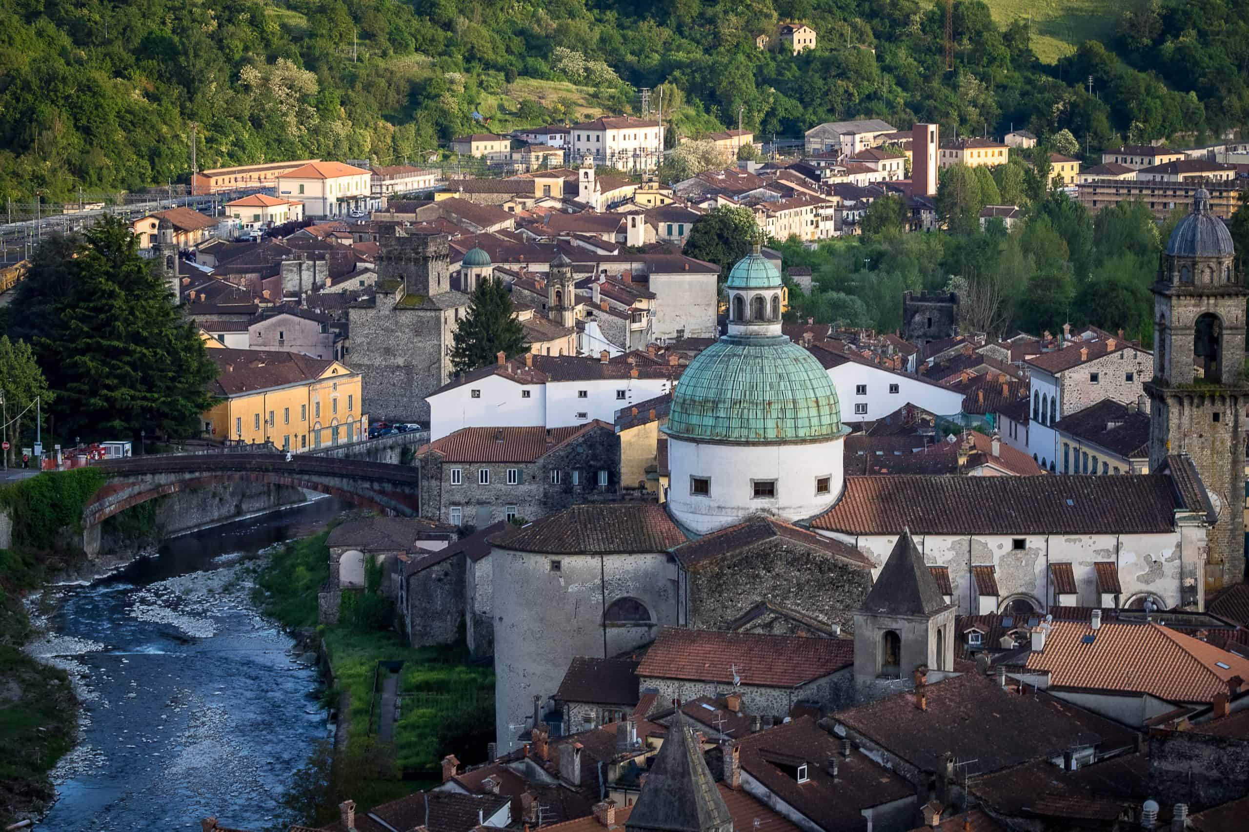 A sweeping view over Pontremoli, Italy, capturing the town’s iconic green-domed church, narrow rooftops, and medieval stone towers. The Magra River curves gently through the scene with a historic bridge spanning it, while golden evening light brushes the surrounding hills and buildings with a warm glow.