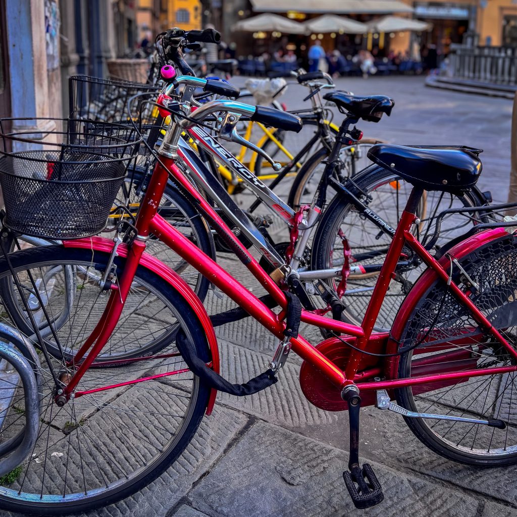 A row of colorful bicycles parked in front of a busy café scene in Lucca, Italy, with locals enjoying drinks under umbrellas near a gelateria.