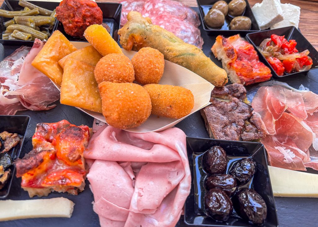 A colorful Italian appetizer spread featuring arancini, fried dough, mortadella, prosciutto, salami, olives, cheeses, and roasted vegetables arranged on a black slate plate with small black dishes.