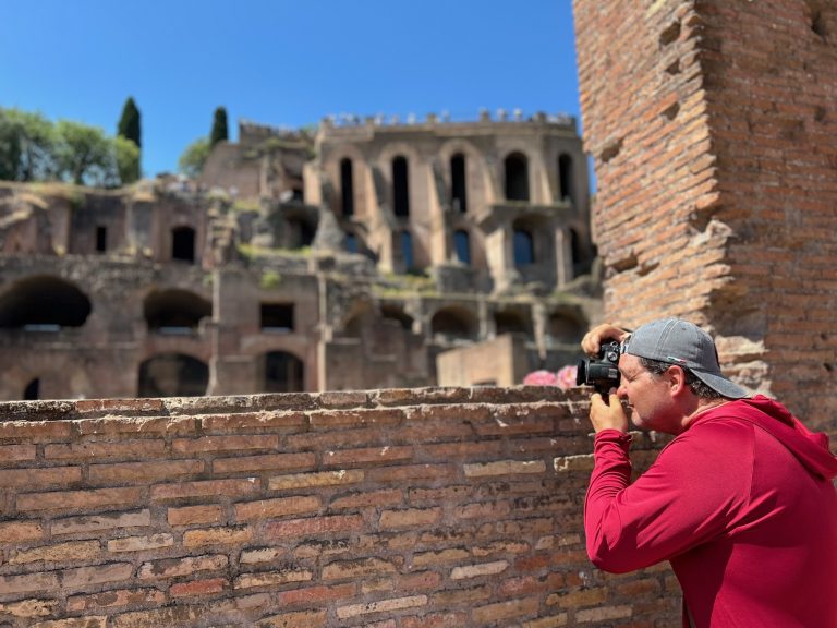 Griffin Smyth in a red hoodie and backwards cap photographs ancient Roman ruins through a brick wall. The background shows weathered stone arches and tall windows under a clear blue sky, with greenery peeking through. The scene blends travel photography with historic architecture.